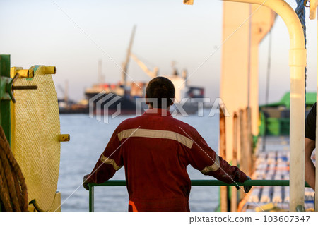 Seaman in red working overall and orange helmet on deck. Working at sea. Cargo vessel. Mooring operations. Able seaman. Seaman in red working overall and orange helmet on deck. Working at sea. Cargo vessel. Mooring operations. Able seaman. 103607347