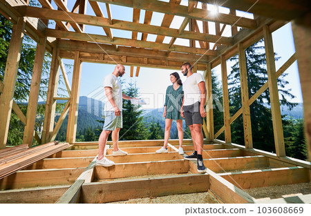 Man with plan showcases to couple the process of constructing wooden framed house. Investors inspecting their future dwelling in mountains near forest. Concept of contemporary ecological construction. Man with plan showcases to couple the process of constructing wooden framed house. Investors inspecting their future dwelling in mountains near forest. Concept of contemporary ecological construction. 103608669