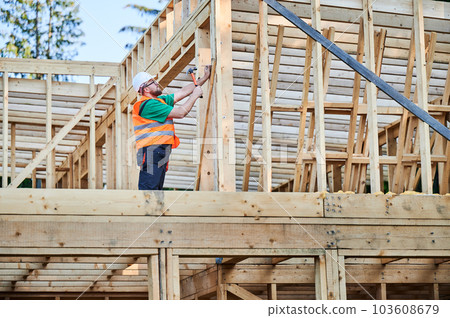 Carpenter constructing two-story wooden frame house near the forest. Bearded man hammering nails into structure while wearing protective helmet and construction vest. Concept of modern construction. 103608679