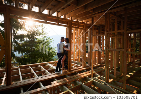 Man and woman examining their future wooden frame dwelling nestled in the mountains near forest. Youthful couple at construction site in early morning. Concept of contemporary ecological construction. 103608701