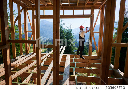 Carpenter constructing wooden frame, two-story house near forest. Bearded man hammering nails with hammer while dressed in protective helmet and work coveralls. Carpenter constructing wooden frame, two-story house near forest. Bearded man hammering nails with hammer while dressed in protective helmet and work coveralls. 103608710