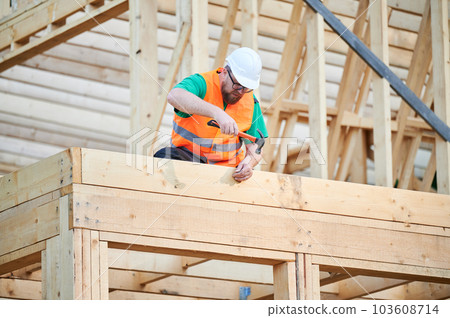 Carpenter constructing two-story wooden frame house. Man wearing glasses hammering nails into structure while wearing protective helmet and construction vest. Concept of modern ecological construction 103608714