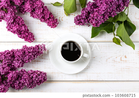 Cup of fresh black coffee and branch of blooming purple lilac with green leaves on white wooden background. Coffee break. Spring flowers. Flat lay, top view, copy space. Cup of fresh black coffee and branch of blooming purple lilac with green leaves on white wooden background. Coffee break. Spring flowers. Flat lay, top view, copy space. 103608715