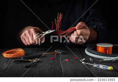 Electrical wires for connecting. Repair of electrical equipment in the workshop of a master electrician. Close-up of the hands of professional master during work. Electrical wires for connecting. Repair of electrical equipment in the workshop of a master electrician. Close-up of the hands of professional master during work. 103613993