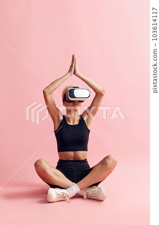 Young woman with slim body, in black sportswear and vr glasses sitting and meditating against pink studio background Young woman with slim body, in black sportswear and vr glasses sitting and meditating against pink studio background 103614117