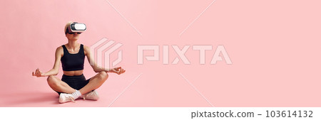 Young woman with slim body, in black sportswear and vr glasses sitting and meditating against pink studio background 103614132
