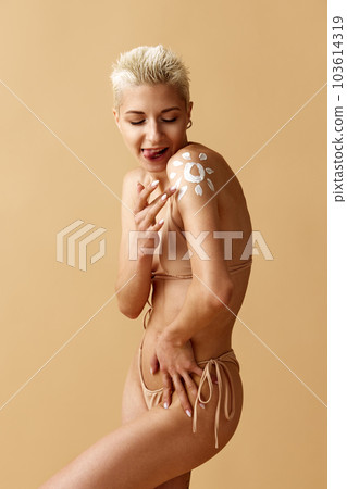 Portrait of beautiful young blonde woman applying spf cream on body, posing in swimsuit against pastel yellow studio background 103614319