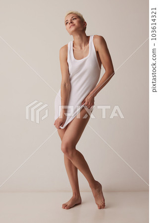 Full-length portrait of slim, beautiful young woman posing in white singlet, homewear against grey studio background 103614321