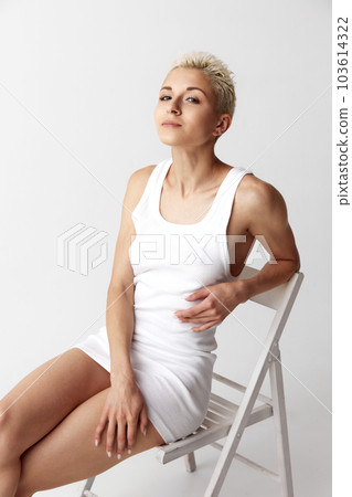 Portrait of beautiful young woman with short blonde hair posing in white singlet, homewear against grey studio background Portrait of beautiful young woman with short blonde hair posing in white singlet, homewear against grey studio background 103614322