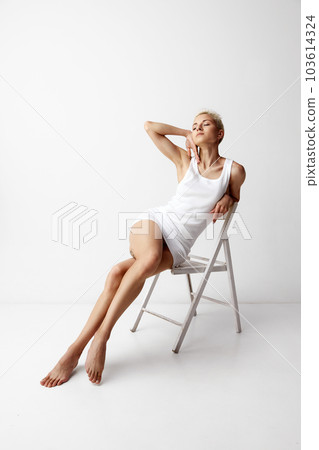 Portrait of tender blonde woman posing on chair in white singlet, homewear against grey studio background Portrait of tender blonde woman posing on chair in white singlet, homewear against grey studio background 103614324