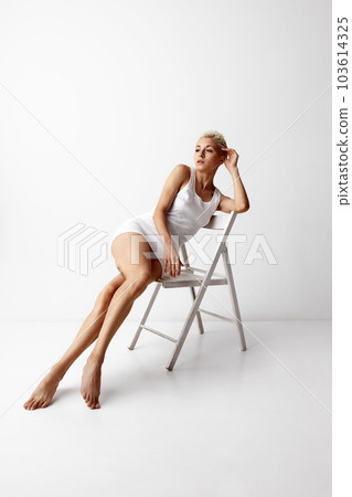 Portrait of tender blonde woman posing on chair in white singlet, homewear against grey studio background 103614325