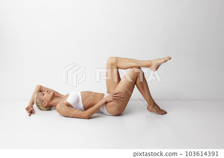 Portrait of blonde young girl with short hair, slim body shape, lying on floor and posing in underwear against grey studio background 103614391