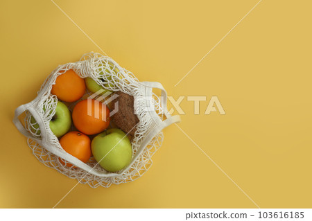 Net bag with fruits on yellow background, top view. Space for text 103616185