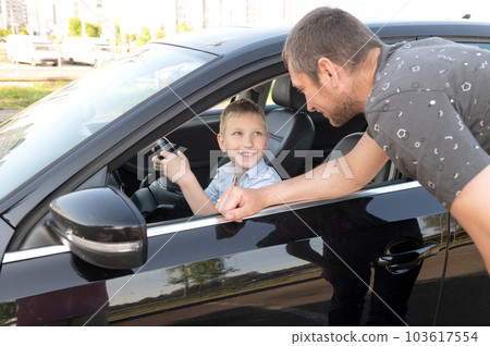 Cute boy is sitting at the wheel in the car, dad is looking out the car window 103617554