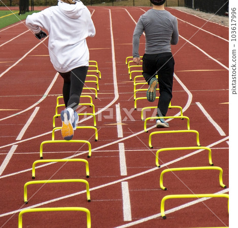 Rear view of a boy and girl track runners running over yellow mini hurdles on a track 103617996