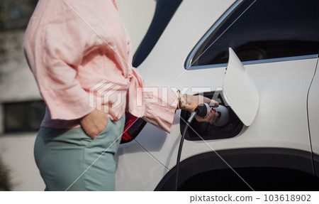 Close-up of woman holding power supply cable from her electric car, charging it, sustainable and economic transportation concept. Close-up of woman holding power supply cable from her electric car, charging it, sustainable and economic transportation concept. 103618902