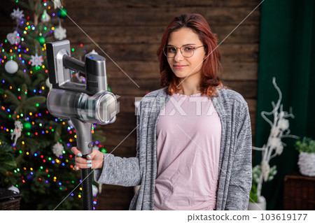The girl is standing, smiling gently against the backdrop of the Christmas tree and holding a vacuum cleaner in her hand. 103619177