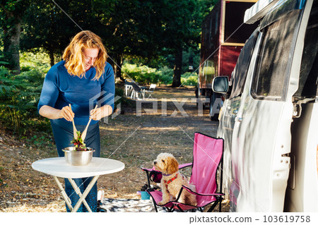 Middle aged nomad woman cooking salad outside her camper van in the forest camp with her cockapoo puppy pet during road trip. Best friend. Enjoying free lifestyle, travel vacation. Travel with pets 103619758