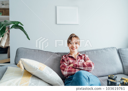 Happy smiling proud of herself young woman relaxing on couch after she puts a picture on the wall at her home. Housekeeping work. Doing repair herself. DIY, gender equality concept. Home decorating 103619761