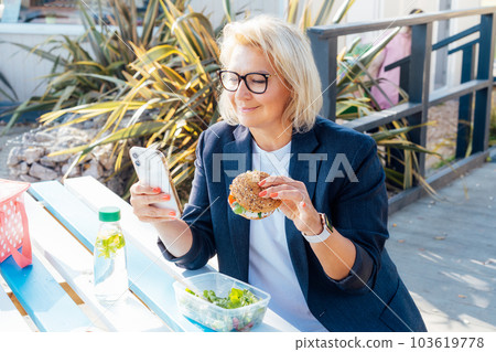 Middle-aged business woman eating a wholemeal burger with salad and using a phone sitting at picnic table in the park while having a break. Healthy lunch box for weight loss. Balanced diet. Middle-aged business woman eating a wholemeal burger with salad and using a phone sitting at picnic table in the park while having a break. Healthy lunch box for weight loss. Balanced diet. 103619778