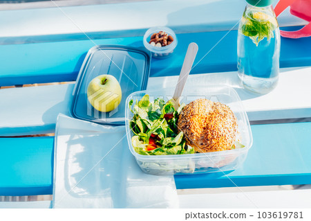 Top view healthy lunch box with wholemeal burger bun, salad with fresh vegetables, a bottle of water with mint and lemon, apple and nuts on the wooden picnic table. Balanced portion with healthy diet. Top view healthy lunch box with wholemeal burger bun, salad with fresh vegetables, a bottle of water with mint and lemon, apple and nuts on the wooden picnic table. Balanced portion with healthy diet. 103619781