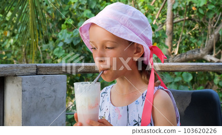 Little girl drinks strawberry milkshake outdoors, resting on a tropical terrace Little girl drinks strawberry milkshake outdoors, resting on a tropical terrace 103620226