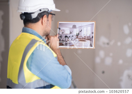 Pensive engineer in protective helmet thinking about Scandinavian interior design while looking at picture in hand against shabby wall in living room renovation process in apartment 103620557