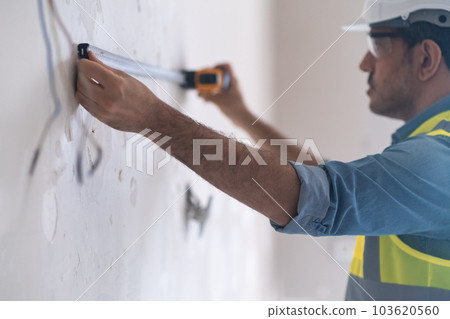Construction worker measuring shabby wall near hanging wires preparing for socket installation focused specialist helping owners with renovation of house interior Construction worker measuring shabby wall near hanging wires preparing for socket installation focused specialist helping owners with renovation of house interior 103620560