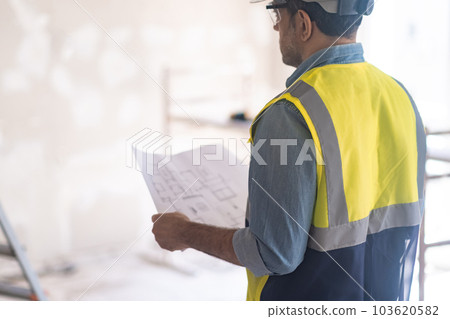 Experienced worker in professional uniform checks condition of wall in room holding project drawing in hands man in vest preparing for renovation process in house 103620582
