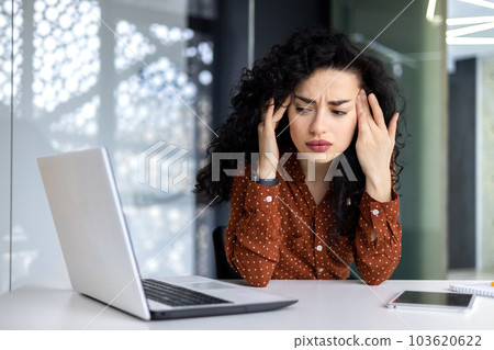 Sick and overtired Latin American business woman working inside office building, female worker holding her head with hands having severe headache and migraine. 103620622