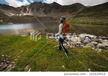 young athletic woman walks through a green field with flowers next to a lake with picturesque mountains. A tourist with a backpack walks with trekking poles on a hike young athletic woman walks through a green field with flowers next to a lake with picturesque mountains. A tourist with a backpack walks with trekking poles on a hike 103620918