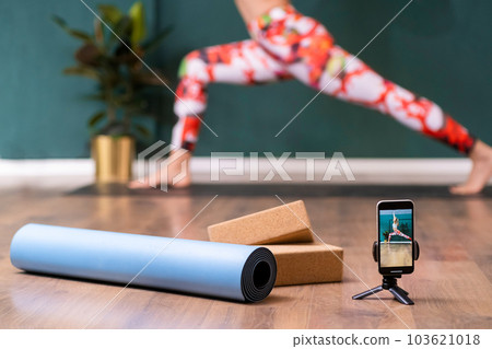 Professional yoga training in cozy studio tripod with phone and mat roll on floor against woman standing in pose female person recording exercise on camera to post on social media 103621018