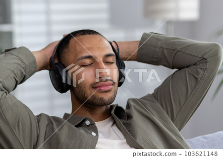 Close-up photo. Portrait of a young African American man listening to music in headphones. Sitting on the sofa with closed eyes, resting. 103621148