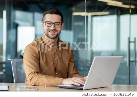 Portrait of a young male office worker, businessman smiling and posing at the camera and working on a laptop while sitting at a desk. 103621149