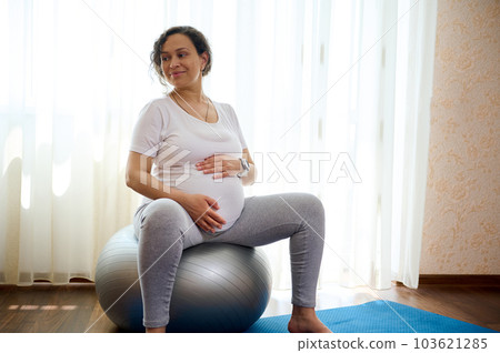 Beautiful curly haired adult pregnant woman holding her belly, sitting on a fitness ball, smiling looking aside, enjoying prenatal fitness at home. The concept of active healthy lifestyle in pregnancy Beautiful curly haired adult pregnant woman holding her belly, sitting on a fitness ball, smiling looking aside, enjoying prenatal fitness at home. The concept of active healthy lifestyle in pregnancy 103621285