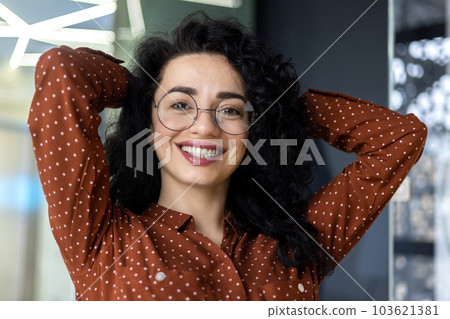 Closeup portrait of successful businesswoman inside office, hispanic woman with curly hair and glasses smiling and looking at camera, boss female with hands behind head satisfied achievements. 103621381