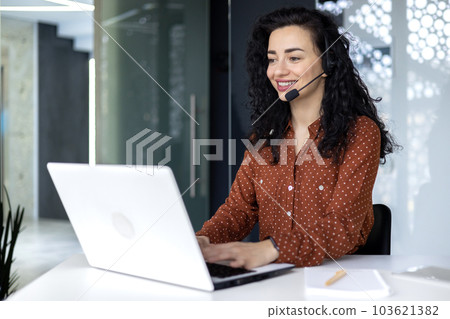 Successful telemarketing and online sales woman working inside office with laptop, mature experienced hispanic woman typing on keyboard, using headset for video call. 103621382