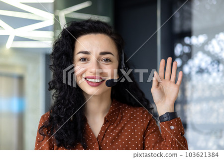 Video call online meeting , businesswoman with headset smiling and looking at web camera, waving hand greeting, mature and experienced hispanic woman working inside office at workplace close up. 103621384