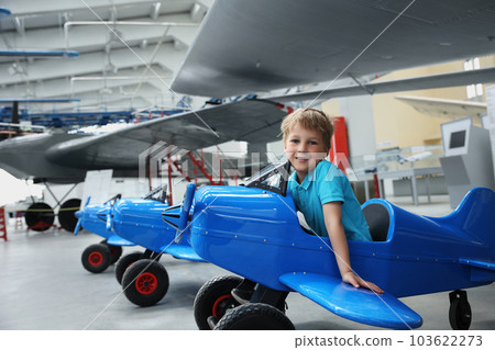 Portrait of cute little blond happy kid boy enjoy have fun play riding pedat toz plane model against vintage airplane background in museum hangar. Future child profession dream concept 103622273