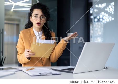 Portrait of disappointed business woman, latin american received notification letter from bank with bad news, office worker looking sad at camera, working at work using laptop. Portrait of disappointed business woman, latin american received notification letter from bank with bad news, office worker looking sad at camera, working at work using laptop. 103622320