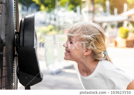 Profile side close-up view cute little blond kid girl enjoy fresh moisturized air blowing from big cooling fan machine hot sunny summer weather outdoors. Mist spraying ventilator system city street Profile side close-up view cute little blond kid girl enjoy fresh moisturized air blowing from big cooling fan machine hot sunny summer weather outdoors. Mist spraying ventilator system city street 103623550