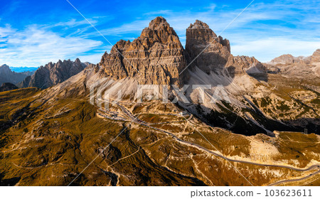 Aerial View of Three peaks of Lavaredo, Alpine Mountain Ranges beneath a Blue Sky with White Clouds, Rocky Peaks Towering over Expansive Shaded Canyon, Bathed in the Warm Glow of Sunset. 103623611