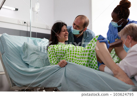 Woman having painful labor, medical team trying to delivery baby during caesarean surgery in hospital ward. Patient with pregnancy pushing for child in maternity clinic being comforting by husband 103624795