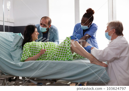 Diverse hospital team helping with child delivery at maternity clinic. Woman lying in bed having painful labor contractions giving birth to baby while husband standing beside her comforting 103624797