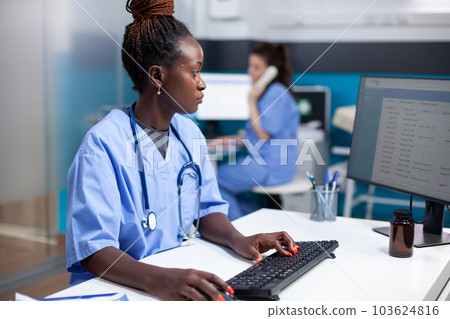 African american nurse working at desk, inserting data in appointment table list. Concentrated adult woman checking patient list, wearing stethoscope in clean, modern medical office operating computer African american nurse working at desk, inserting data in appointment table list. Concentrated adult woman checking patient list, wearing stethoscope in clean, modern medical office operating computer 103624816