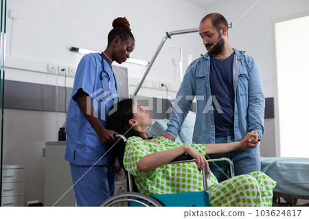 African american nurse transporting pregnant woman in wheelchair to delivery room. Patient with with baby bump preparing for childbirth while being comforting by husband in hospital ward African american nurse transporting pregnant woman in wheelchair to delivery room. Patient with with baby bump preparing for childbirth while being comforting by husband in hospital ward 103624817
