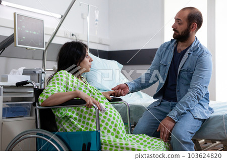 Future father comforting mother who sitting in wheelchair, discussing about child delivery before caesarean surgery. Young parents waiting for childbirth in hospital ward. Medicine concept 103624820
