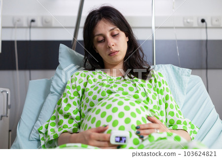 Pregnant woman holding hand on belly, lying in bed preparing for childbirth in hospital ward. Caucasian patient with pregnancy wearing oximeter, expecting child in maternity clinic 103624821