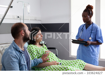 African american assistant discussing childbirth process with pregnant woman, preparing mother for surgery in hospital ward. Patient with pregnancy lying in bed waiting for gynecologist doctor 103624827