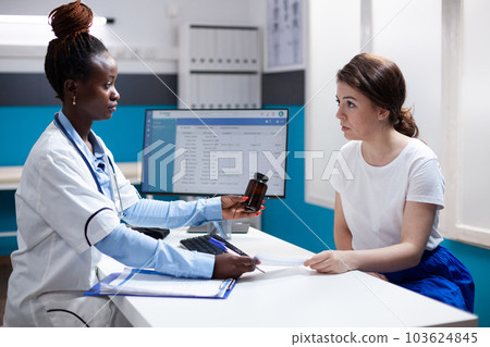 African american doctor prescribing pill bottle medication to patient. Healthcare specialist during medical checkup in clean modern hospital office offering diagnosis to woman. 103624845